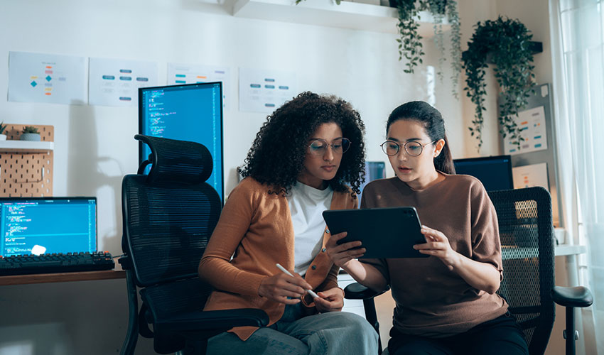 Women in tech collaborate on UI review with tablet and coding monitors in background