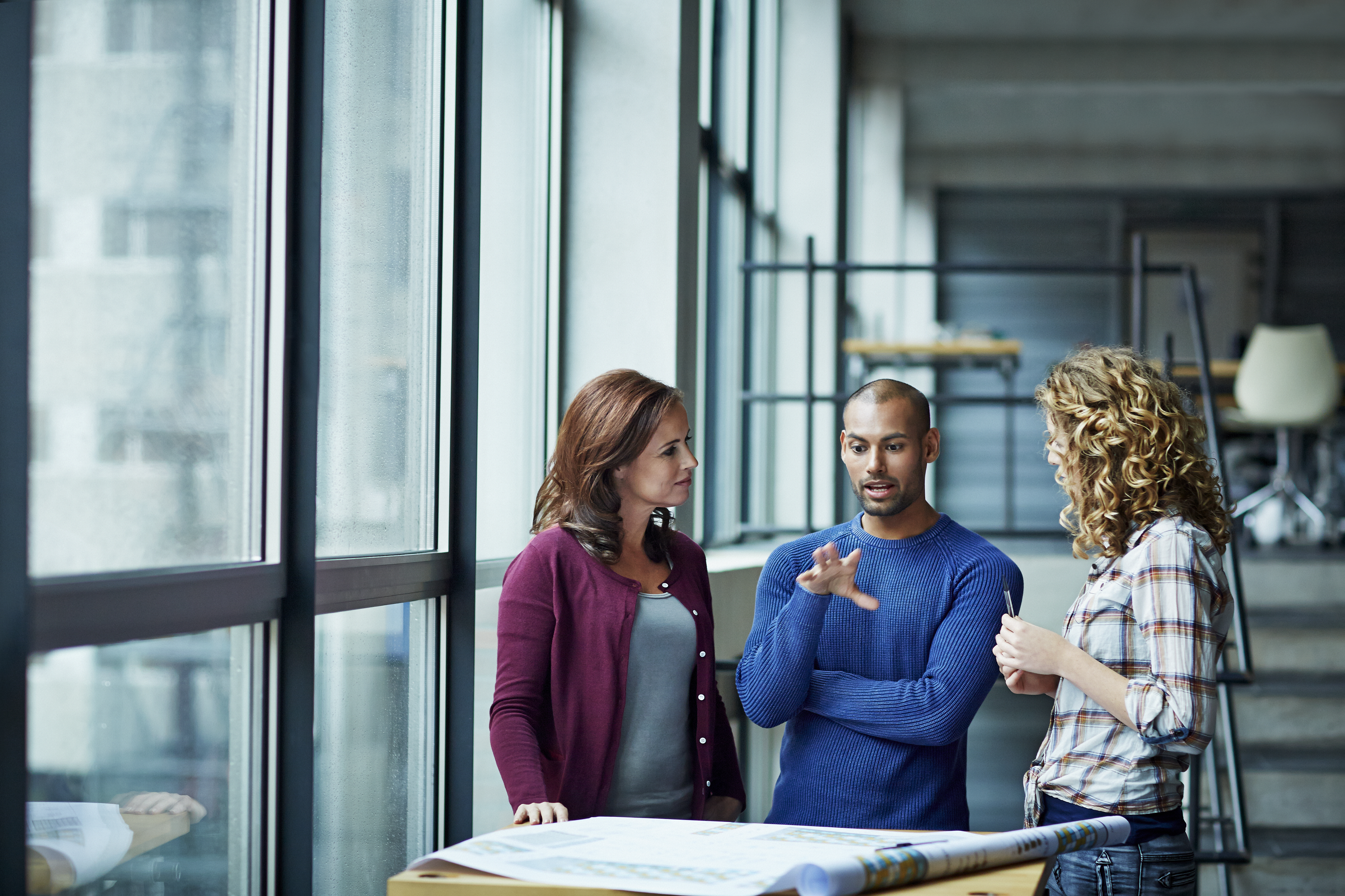 Three colleagues discuss architectural plans in a bright, modern office by a window