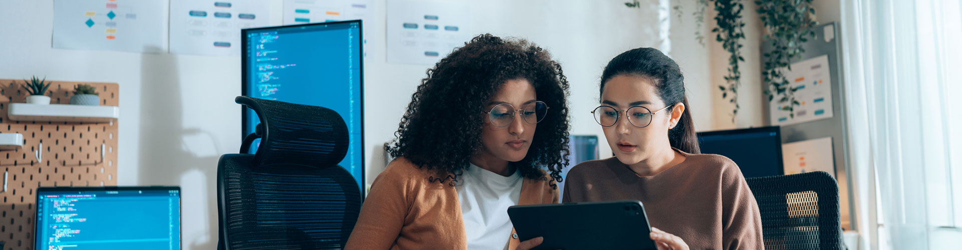 Women in tech collaborate on UI review with tablet and coding monitors in background