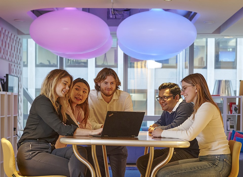Five colleagues look at a laptop in a relaxed office environment