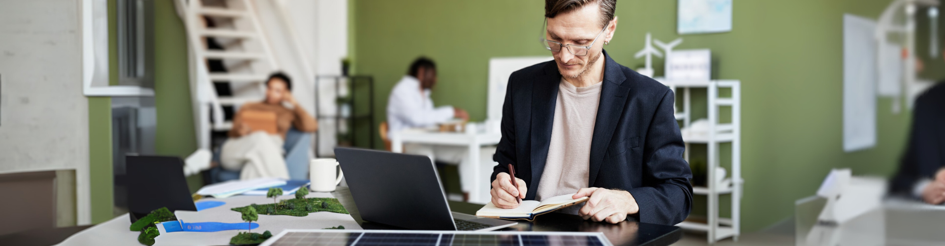 Young businessman making notes in notebook while working at table with laptop and solar panel