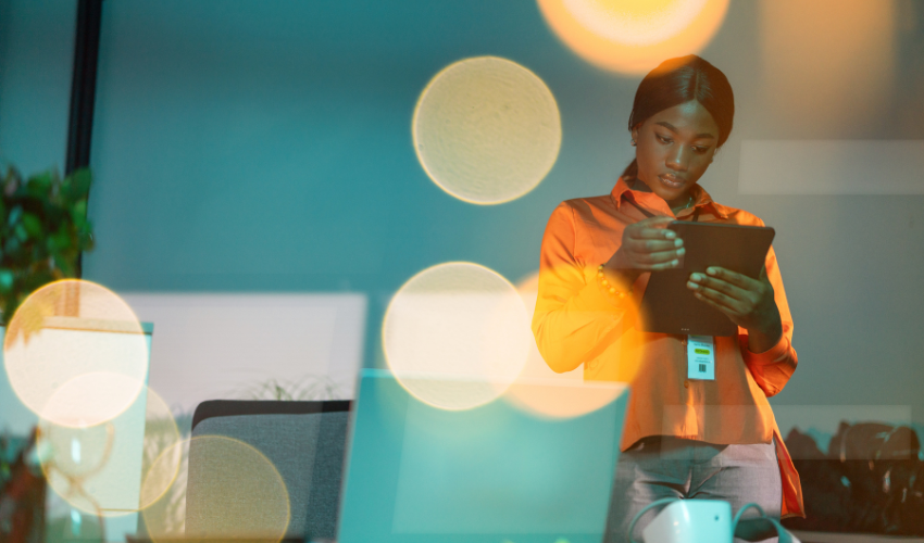 Portrait of young motivated businesswoman surrounded with bokeh lighting standing by the desk and using her digital tablet while working late in modern office