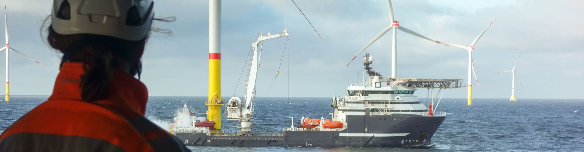 Engineer in high-visibility gear looks out at an offshore wind farm and service vessel
