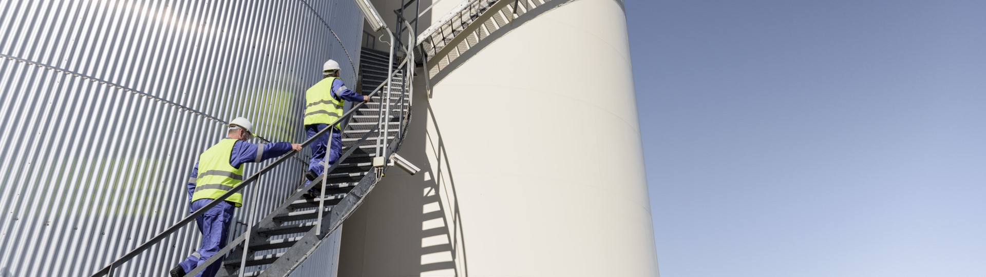 Two men in overalls and hard hats climbing up stairs in a oil storage facility