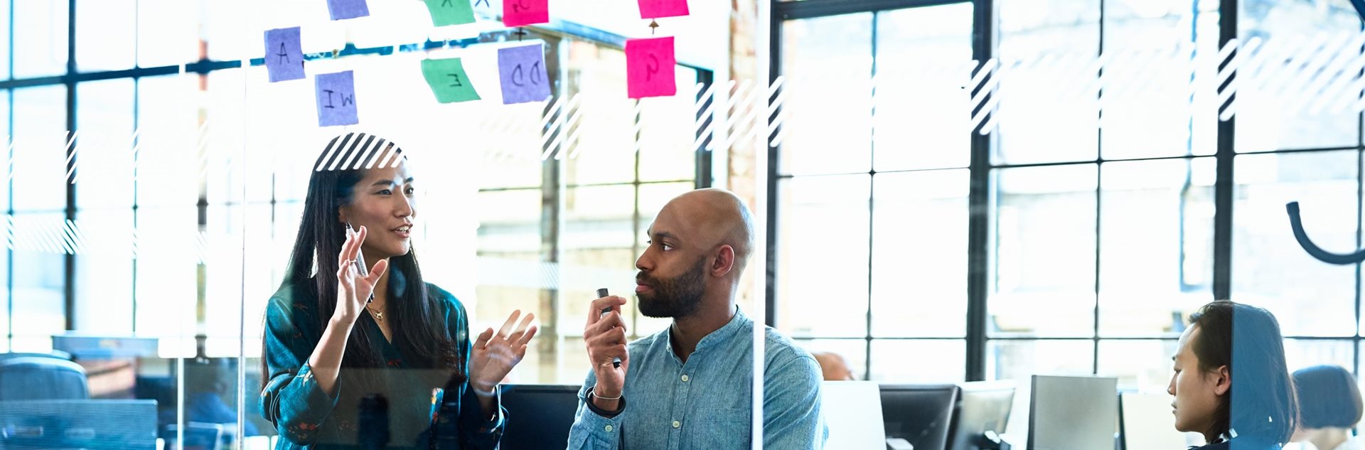 A man and woman discussing post-its on a transparent office wall