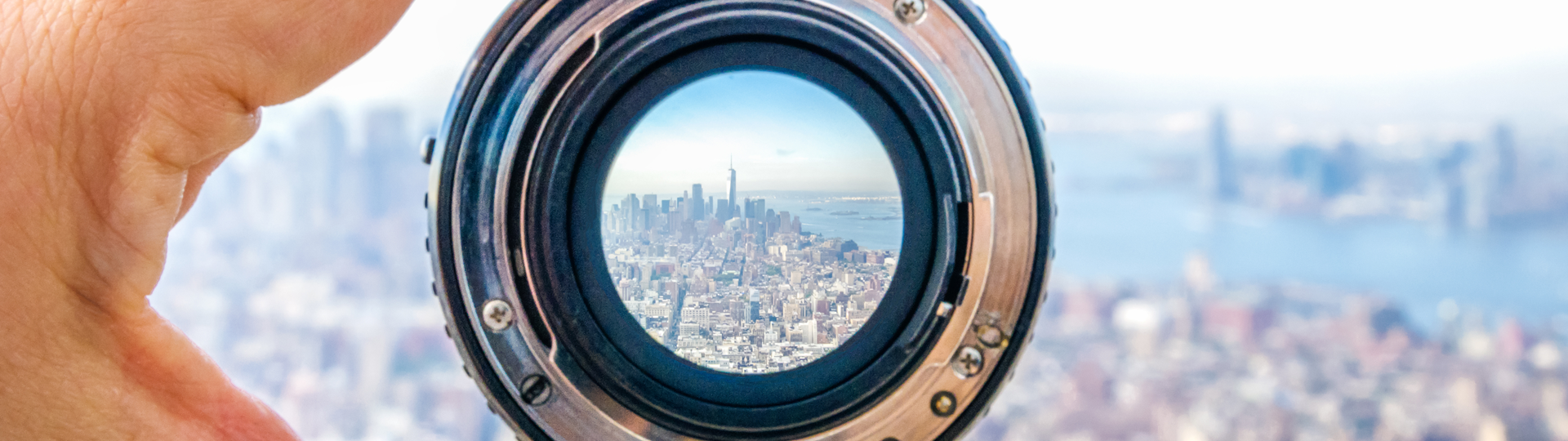 A hand holding a camera lens with a view the Lower Manhattan cityscape