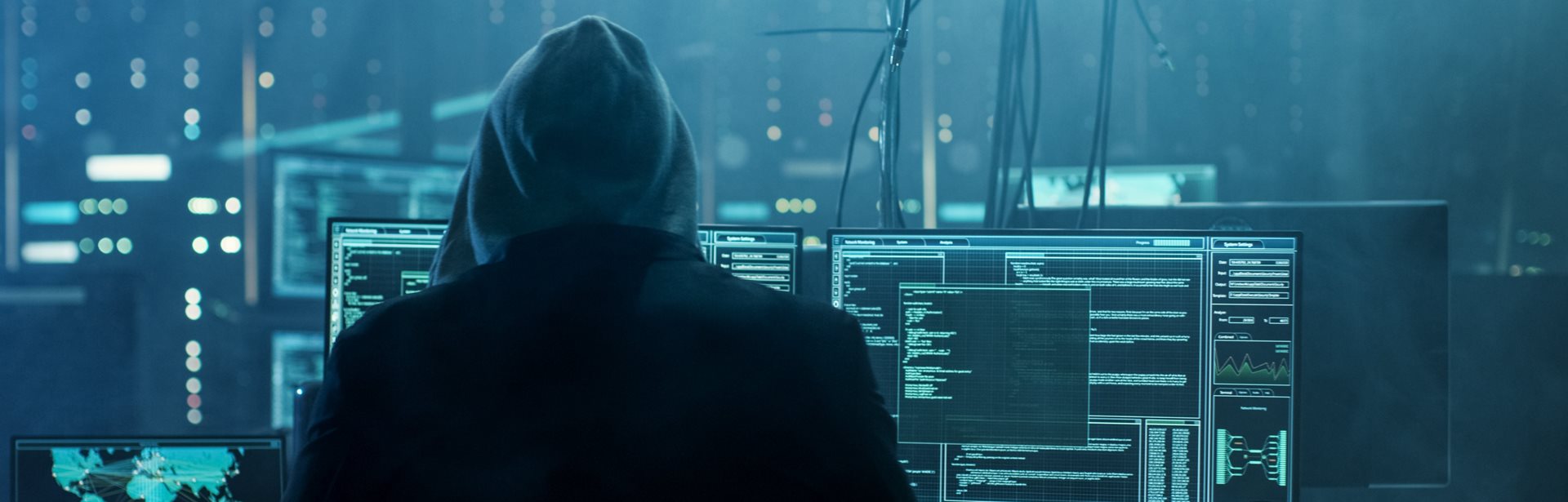 A hacker whose identity is hidden by a hooded top looking at multiple screens