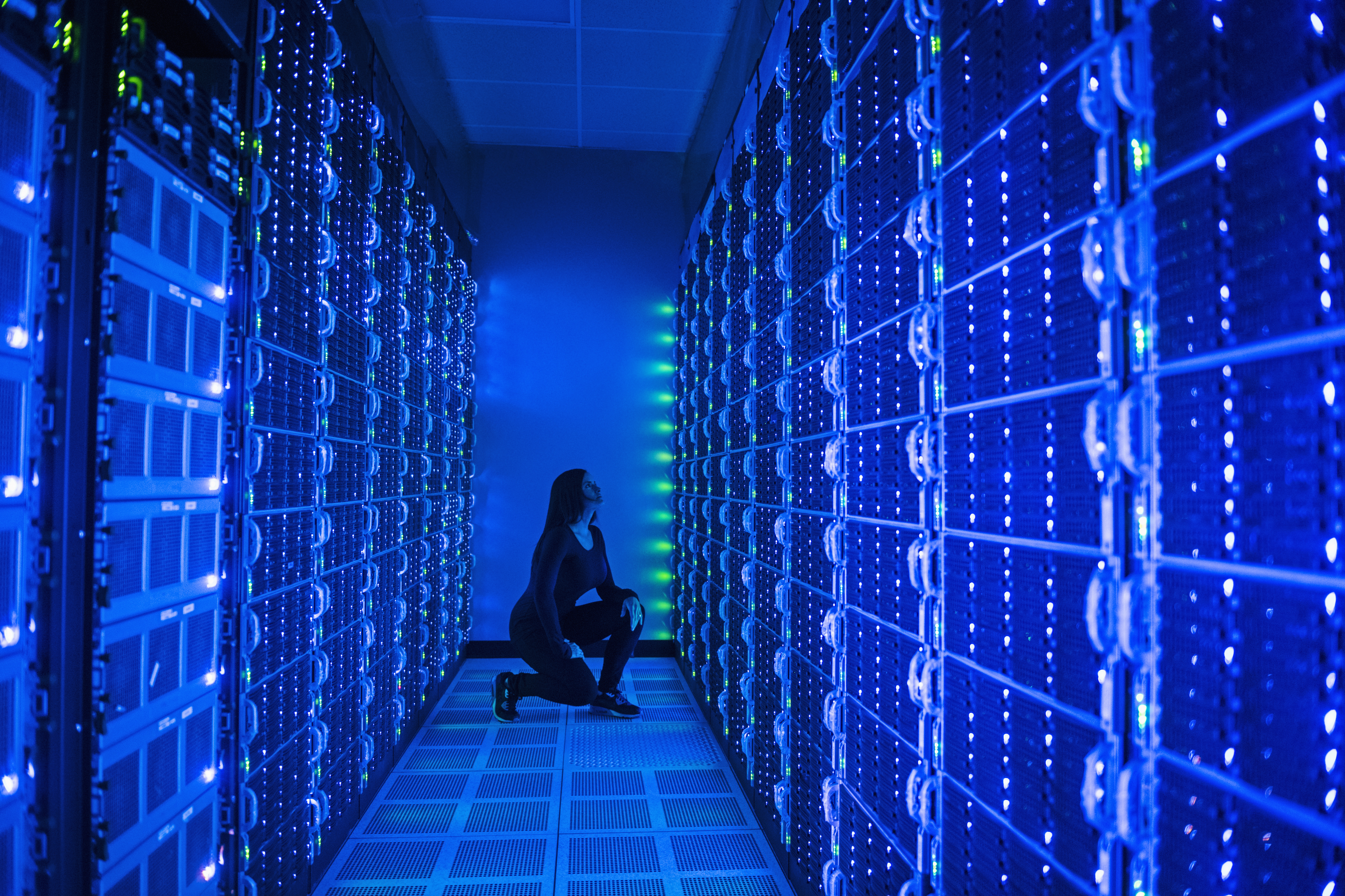 A technician kneels between tall rows of glowing blue server racks in a darkened data center
