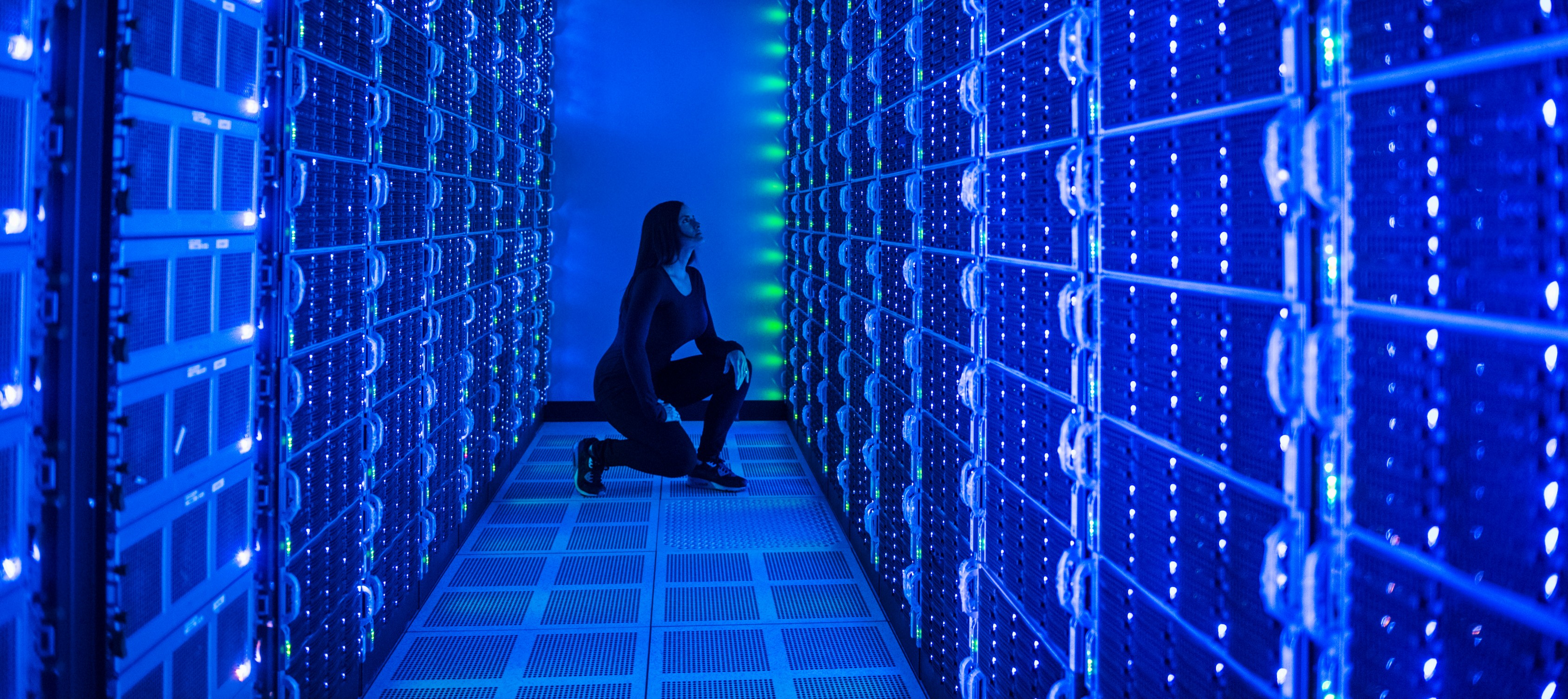 A technician kneels between tall rows of glowing blue server racks in a darkened data center