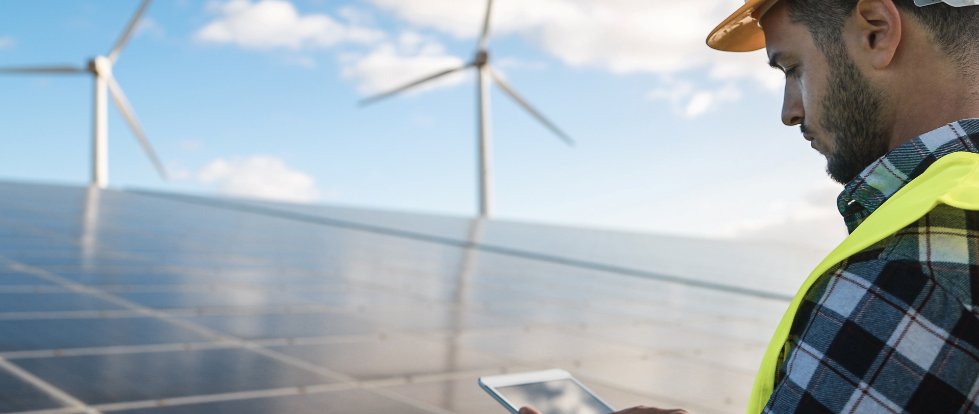 Engineer in hard hat and safety vest using a tablet at a solar and wind farm.