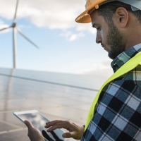 Engineer in hard hat and safety vest using a tablet at a solar and wind farm.