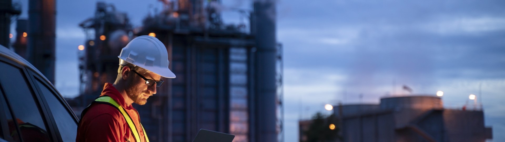 A man in hard hat looking at a tablet in front of industrial buildings at night