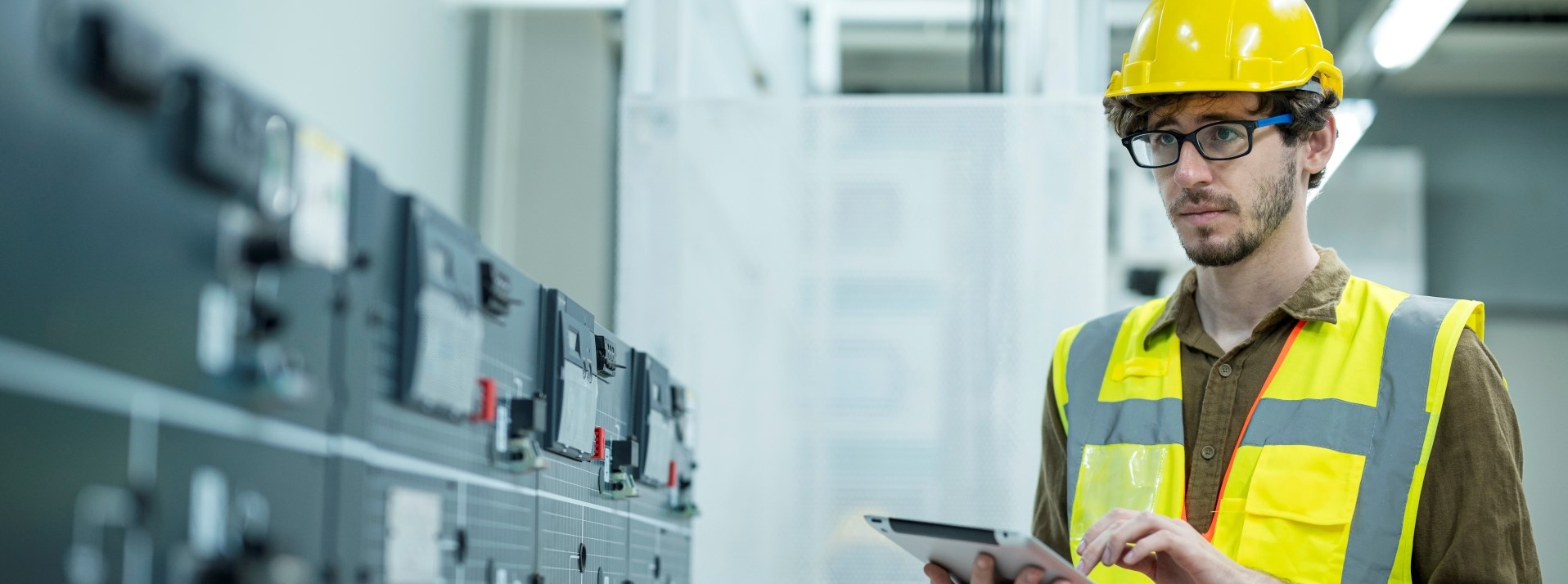 A man with hard hat and glasses holding a tablet while inspecting equipment