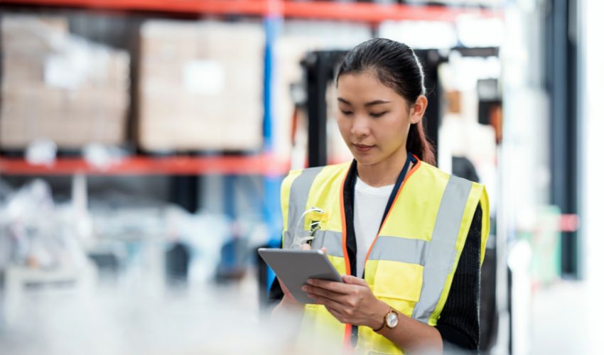 Woman in a high-visibility safety vest using a tablet in a large, blurred warehouse