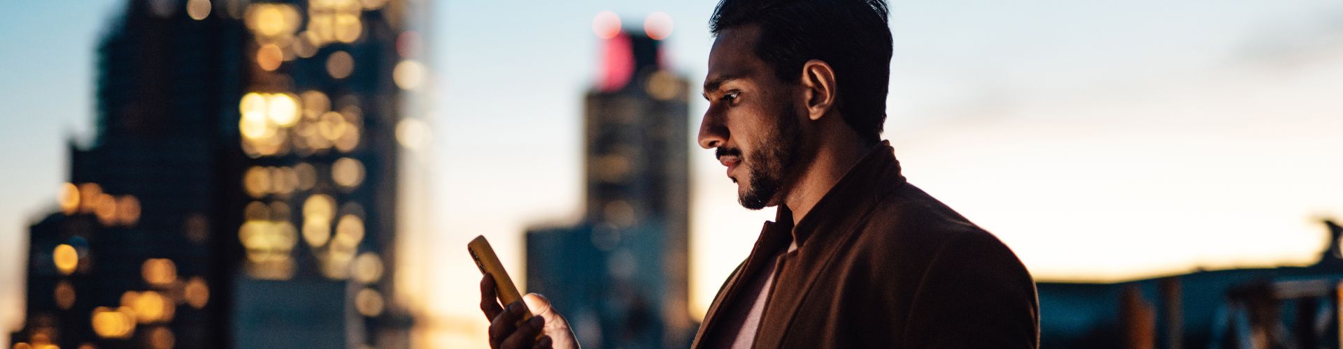 A man checks his smartphone at dusk against a blurred city skyline