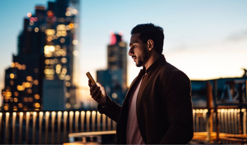 A man checks his smartphone at dusk against a blurred city skyline