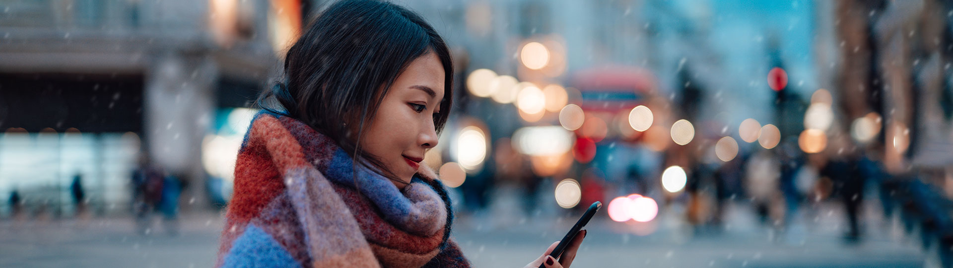 A woman wearing a scarf looking at her phone standing outside while sleet is falling