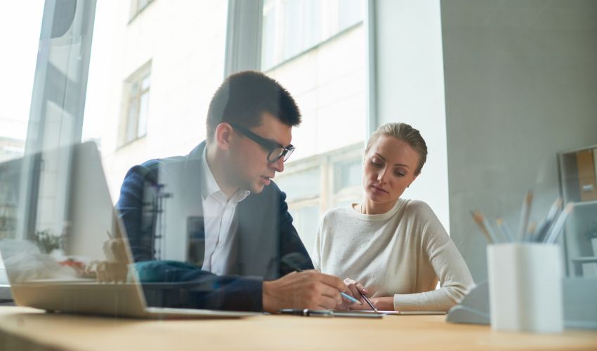 Two colleagues in professional attire discussing documents at a desk in a bright office