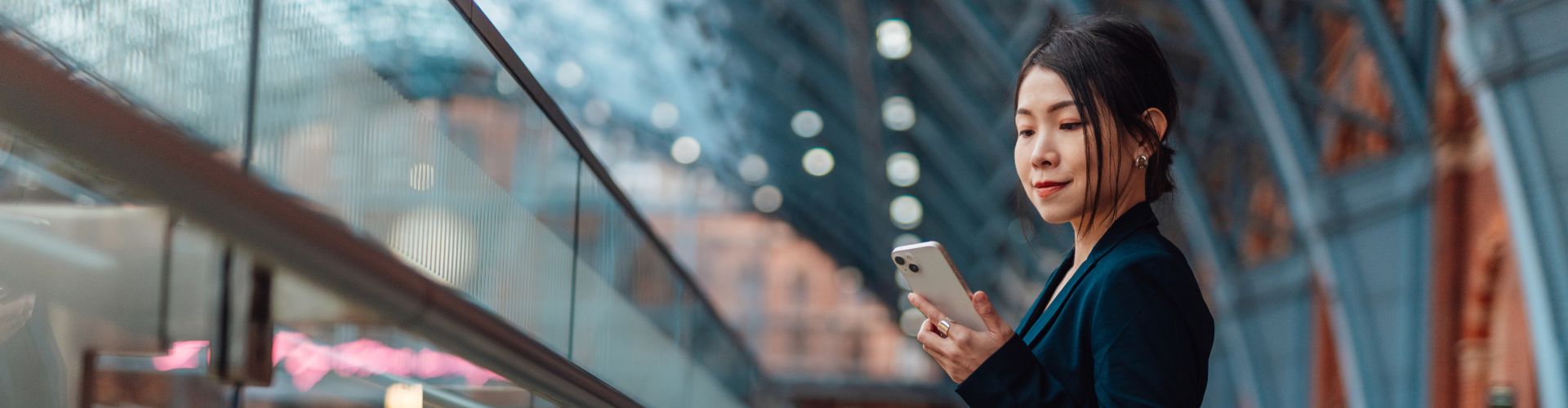 A woman in professional attire using a smartphone in a modern, glass-roofed station