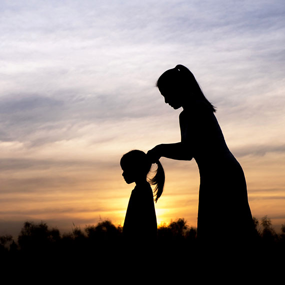 A mum putting her daughter's hair in a pony-tail silhouetted by the setting sun