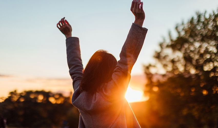 Rear view of young woman stretching arms against sunset in the sky